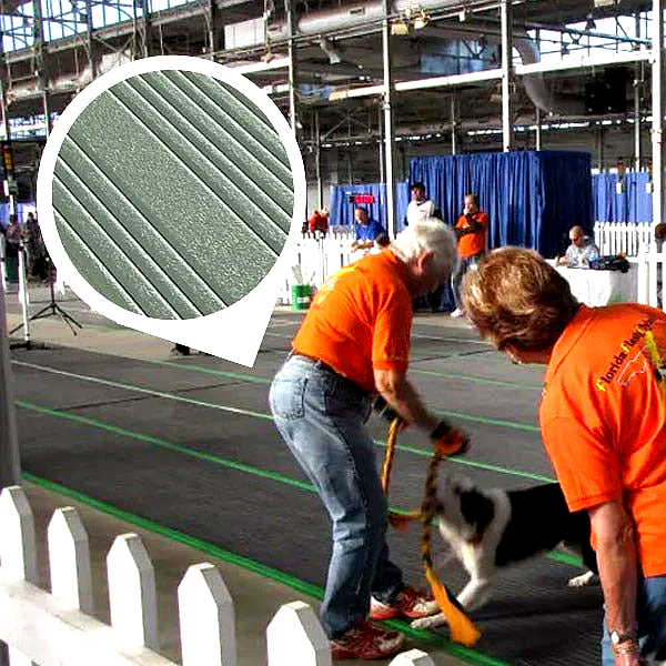 Dark grey anti-fatigue noise reducing matting with a ribbed non-slip surface, providing cushioned support for a large indoor event space. The durable mat features green lane markings, visible as a man and woman train a dog.