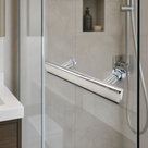 Chrome T-bar handle on a glass shower door, beige tiles with a lit niche and basin unit behind