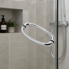 Chrome oval pull handle on a shower screen, beige tiled niche with a plant and shower hose behind
