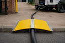 Hi-vis yellow ramp with galvanised chequer-plate spine spanning a black hose at a kerbside, generator trailer behind