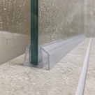 Clear seal at the foot of a wet glass panel on a beige stone tray, flat flange running away along the ledge
