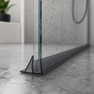 Black seal running along the base of a wet glass shower panel on dark grey tiles, shower hose behind