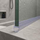 Clear bulb seal at the foot of a wet glass shower panel on a grey stone step, shower hose behind