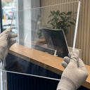 Gloved hands holding a clear acrylic glazing panel at a reception desk, monitor and plant behind