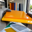 Gloved hands holding stacked gloss acrylic sheets, orange over yellow, samples behind
