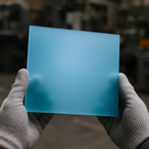 Gloved hands holding a frosted light blue acrylic square, blurred workshop machinery behind
