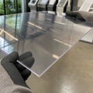 Gloved hands holding a thin clear acrylic sheet in an office foyer, printers and glazing reflected