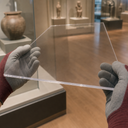 Gloved hands holding a thin clear acrylic panel in a museum gallery, artefact cases behind