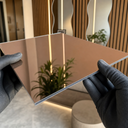 Gloved hands holding a bronze-tinted acrylic mirror sheet reflecting a lit interior