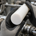 Gloved hands holding a slim white acetal rod, gears and machinery blurred behind
