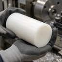 Grey-gloved hands holding a white acetal billet in front of a lathe chuck and swarf