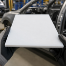 Grey-gloved hands holding a thin white acetal panel in a vehicle workshop, car body shells behind