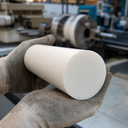 Gloved hands cradling a large cream-white acetal billet, lathe blurred behind in the workshop