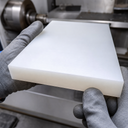 Gloved hands holding a thick milky white nylon block in front of a metalworking lathe