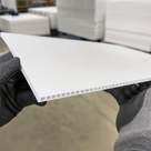 Thin white corrugated sheet held edge-on in gloves, fluted core visible, pallet stacks behind