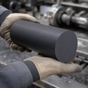 Leather-gloved hands holding a matt charcoal nylon billet, workshop machinery blurred behind
