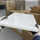 Two gloved hands holding a white polypropylene panel angled up, cardboard boxes and stock behind