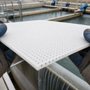 Gloved hands tilting a white perforated sheet above water treatment tanks, round holes in a regular grid