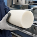 Gloved hand holding a large cream polypropylene billet, factory machinery blurred behind