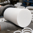 Gloved hands holding a large white PTFE billet, stacks of white PTFE rings behind