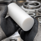 Black-gloved hands holding a white PTFE rod, steel bearing races stacked behind