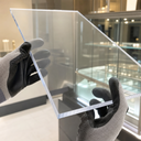 Grey-gloved hands tilting a clear acrylic sheet in a jewellery showroom, lit glass cabinets behind