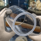 Clear acrylic tube braced in tan gloves, wide sawn rim towards the lens, mineral display cases behind