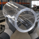 Gloved hands holding a very large-bore clear acrylic tube in a warehouse, metal racking and stock behind.
