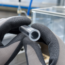 Gloved hands holding a small-diameter clear acrylic tube, cut end to camera, glass tank in the background.