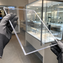 Dark-gloved hands holding a clear acrylic sheet in a showroom, glazed display cabinets seen through it