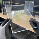 Gloved hands holding a clear acrylic panel above a bench of acrylic sign holders and stands
