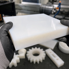 Black-gloved hands holding a thick translucent white PETP slab above machined white gears and brackets