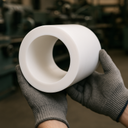 Grey-gloved hands holding a thick-walled white PTFE tube with the open bore forward, blurred machine shop behind