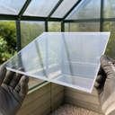 Gloved hands holding a clear acrylic panel in front of a green-framed greenhouse