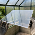 Gloved hands holding a clear acrylic panel in front of a green-framed greenhouse