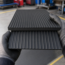 Gloved hands holding two black wide-ribbed rubber mat samples, broad raised ribs and a plain underside
