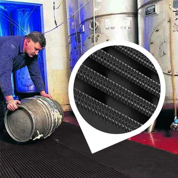 Worker rolling a wet metal barrel (keg) across a black, heavy-duty industrial matting roll in a facility with large storage tanks. The mat is visible in a wet area with a red floor. An inset circle magnifies the mat's non-slip, high-traction surface featuring UV-resistant diamond-patterned ribs, confirming its durability for wet areas, industrial, and outdoor use.