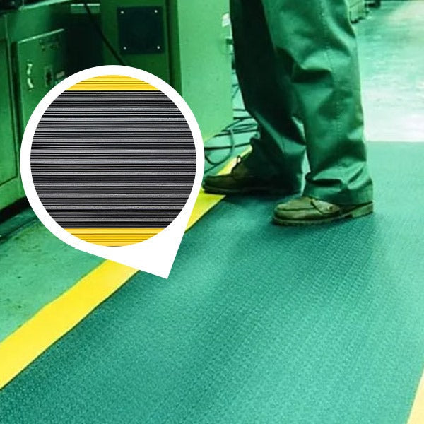 Worker standing on a green anti-fatigue noise reducing mat with a subtle non-slip texture in a factory. The durable PVC foam cushioned mat features high-visibility yellow safety borders, providing cushioned support at an industrial workstation.