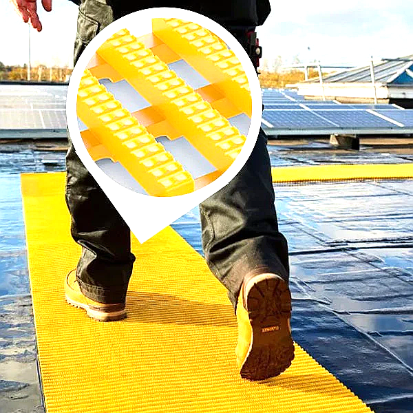 Yellow non-slip safety walkway matting with an integrated drainage system laid on a commercial roof. A worker's boots are seen walking across the highly visible yellow mat, which provides a safety walkway on the dark roof membrane near solar panels. An inset highlights the matting's elevated, ribbed, non-slip surface.