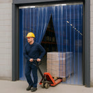 Worker in a yellow hard hat pulls a loaded pallet truck through a blue-tinted PVC strip curtain doorway