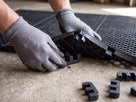 Gloved hands pressing a dual-purpose connector clip into the underside of two open-grid mats to join them