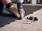 Black matting floor hooks being screw-fixed through an open-rib mat into a concrete workshop floor