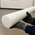 Grey-gloved hands supporting a long white acetal rod indoors, broad round cut end towards the camera
