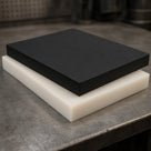 Black acetal slab stacked on a natural-white slab on a steel workshop bench, thick sawn edges