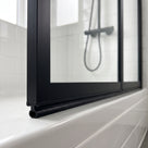 Black oval-tube seal under the bottom rail of a black-framed shower screen, resting on a white tiled bath edge
