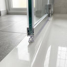 Clear bubble seal running along the foot of a glass shower door on a white tray, chrome hinges beyond