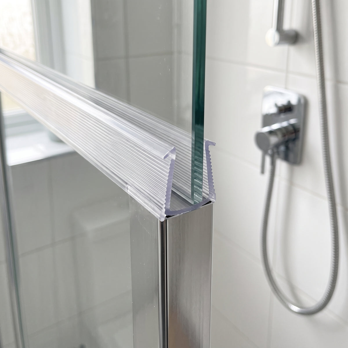 Clear ribbed frame seal on the top rail of a glass shower screen, chrome shower valve and hose behind
