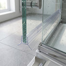 Clear F-shaped channel seal down a glass door edge above a chrome-framed tray, window and grey tiles behind