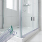 Cut sample of the clear twin-channel seal on a glass offcut, with a shower enclosure corner and white tray behind