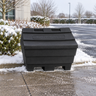Black grit bin with GRIT SALT moulded into its sloped lid, on a snow-edged pavement beside a car park